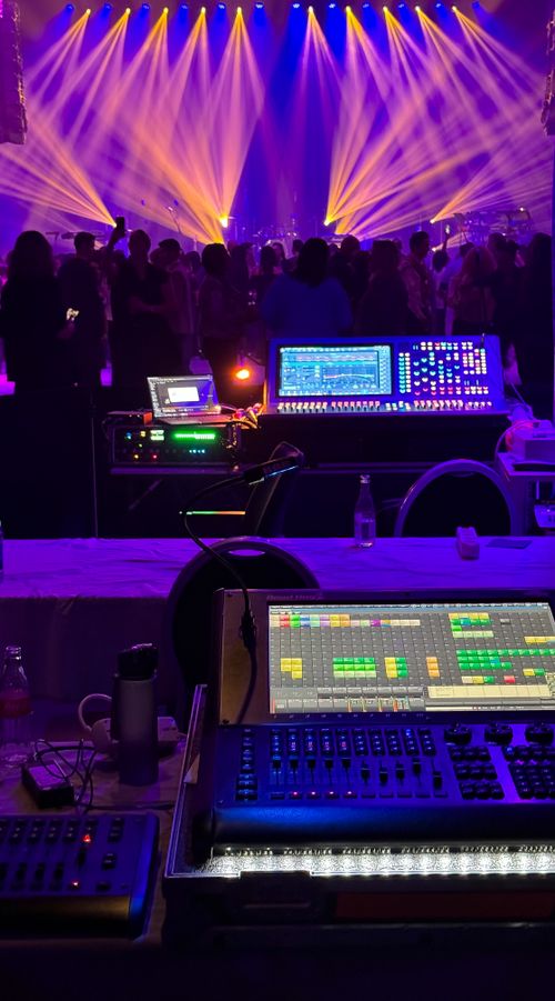 Vue de console et régie lumière au premier plan, avec des panneaux et écrans lumineux et une scène animée par des faisceaux