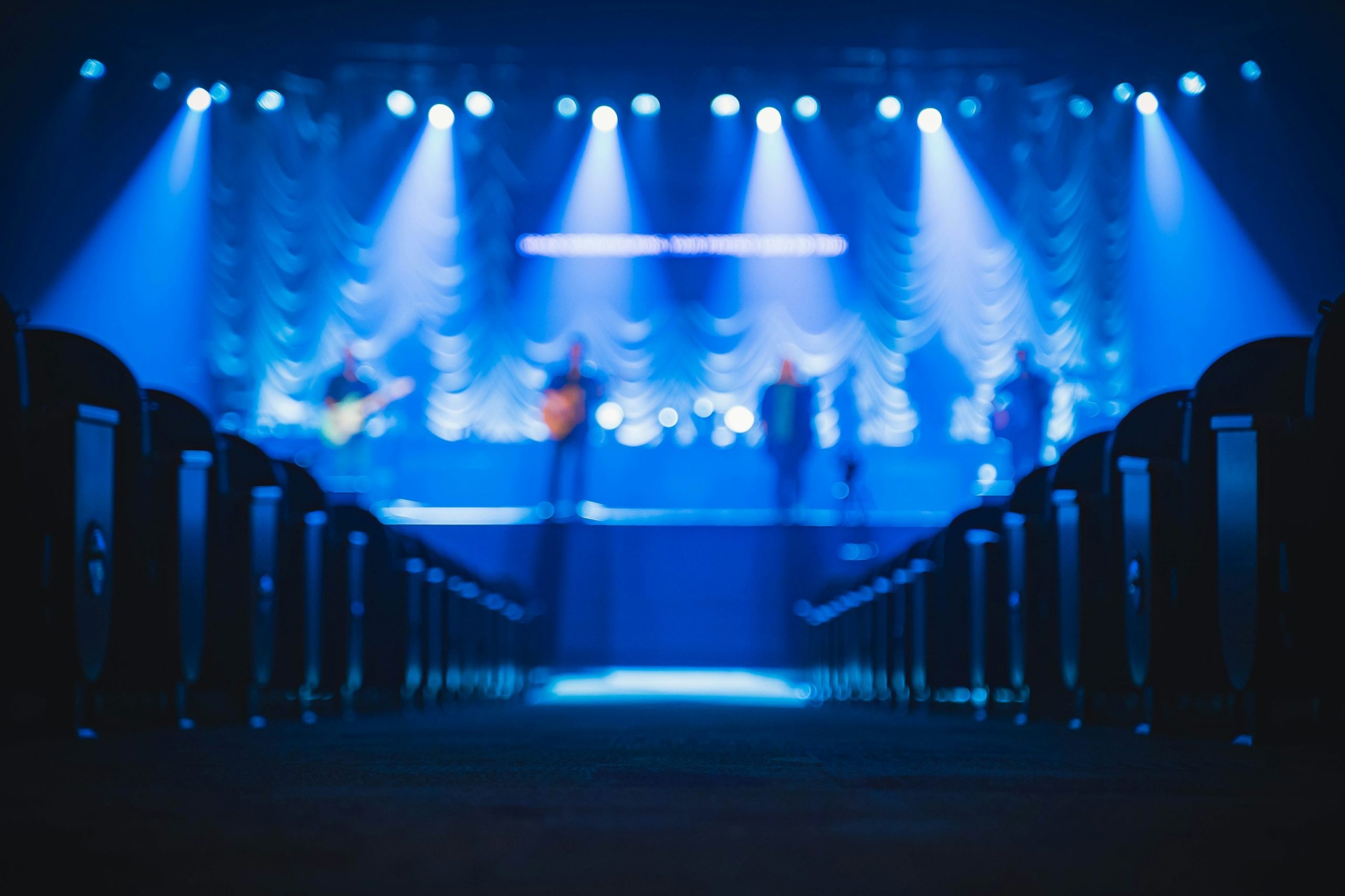 Vue de l’intérieur d’une salle de concert, avec une scène éclairée en bleu et des musiciens flous au fond.